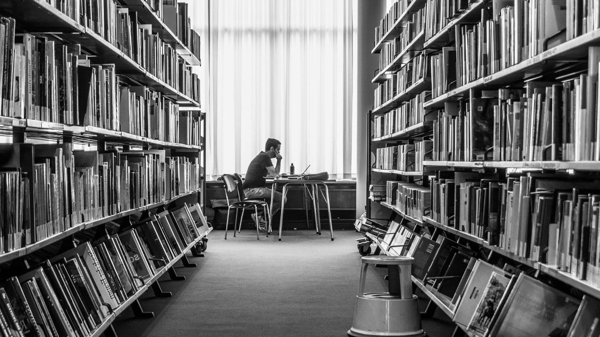 En person sitter vid ett bord med en laptop mellan två bokhyllor i ett bibliotek.