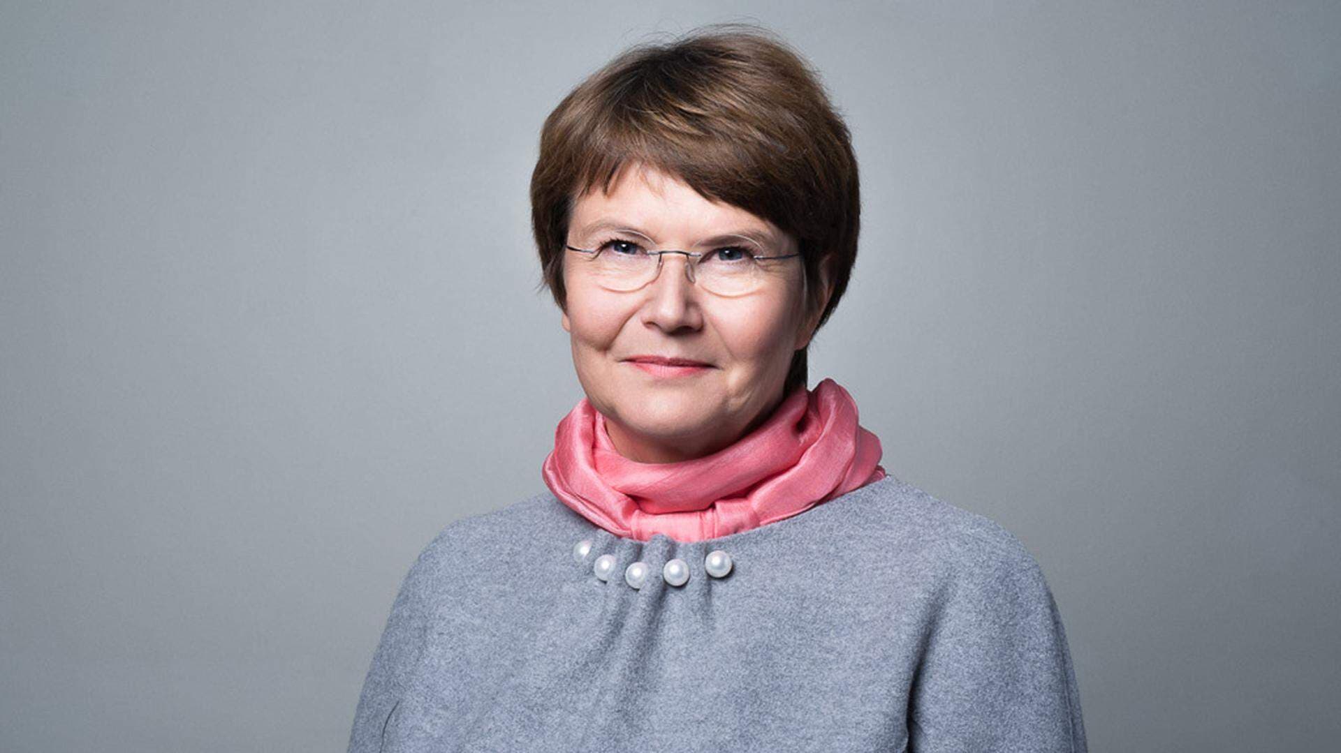 Tuula Teeri i grå tröja och rosa scarf med grå bakgrund