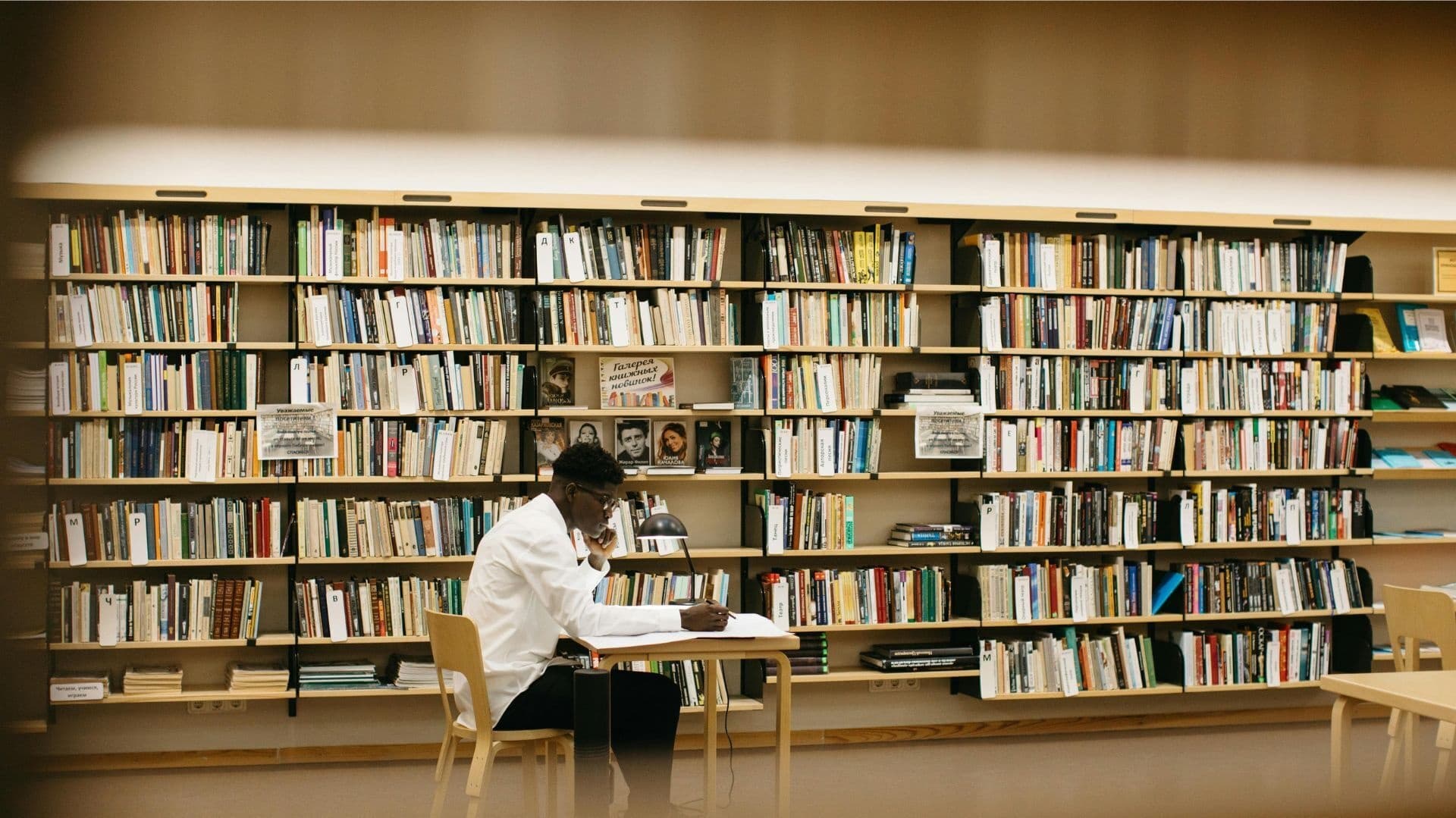 En person sitter och skriver vid ett bord i ett bibliotek, omgiven av fyllda bokhyllor.