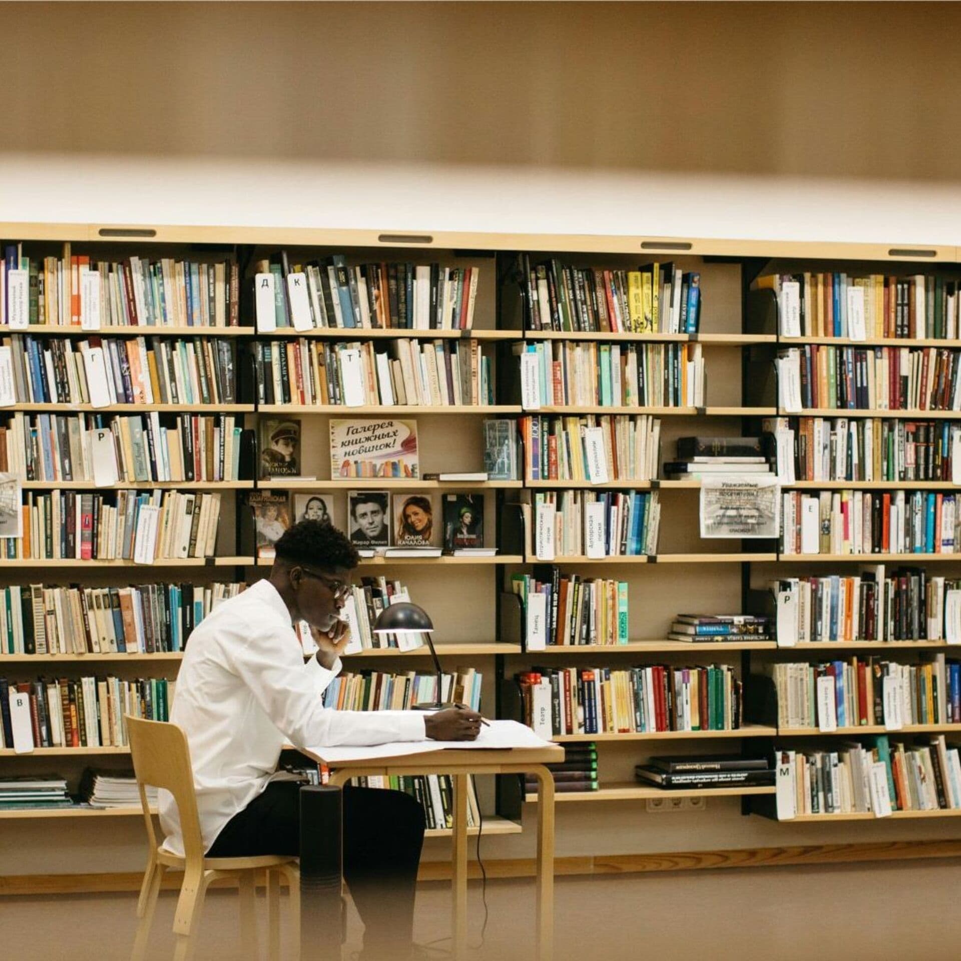 En person sitter och skriver vid ett bord i ett bibliotek, omgiven av fyllda bokhyllor.