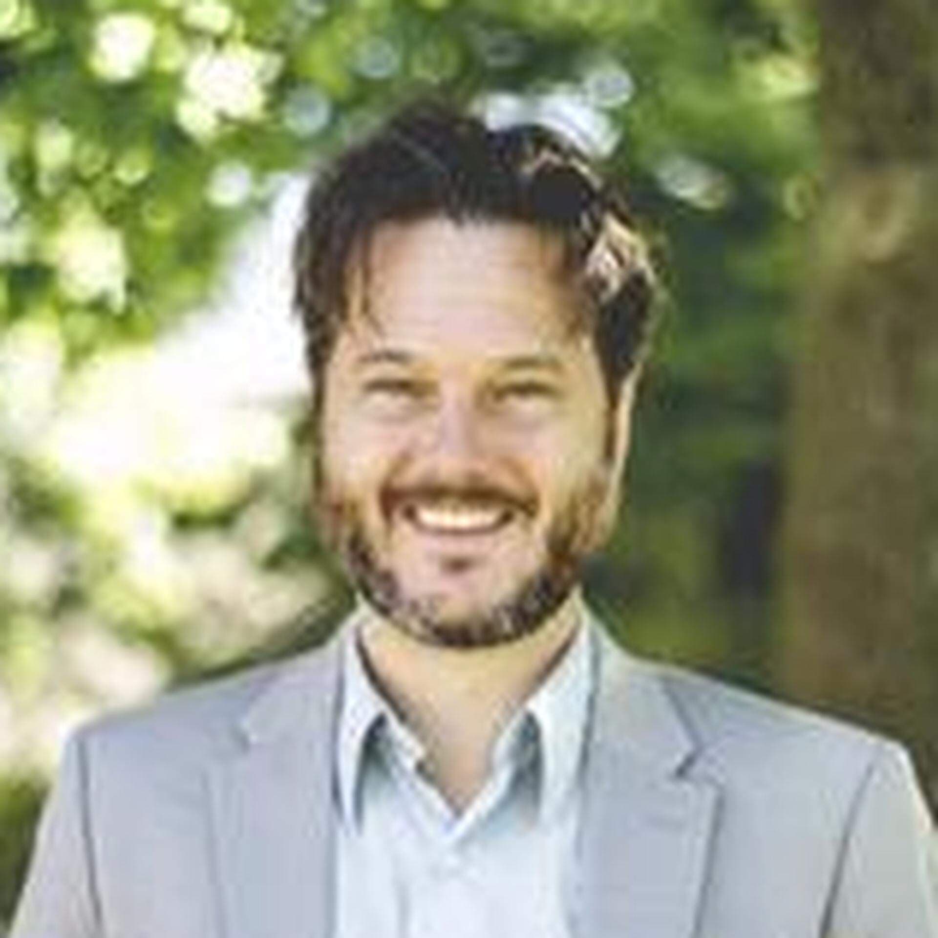 Joakim, iklädd vit skjorta och ljusgrå kavaj, med grönska i bakgrunden