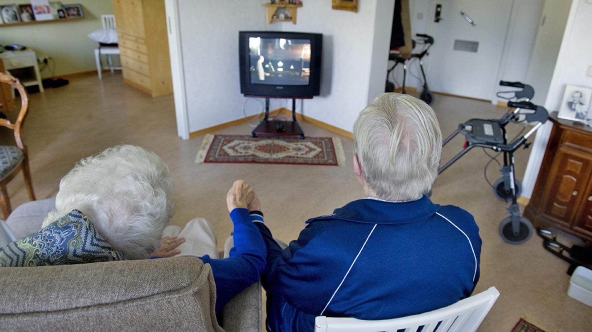 Två pensionärer håller varandra i handen och tittar på tv.