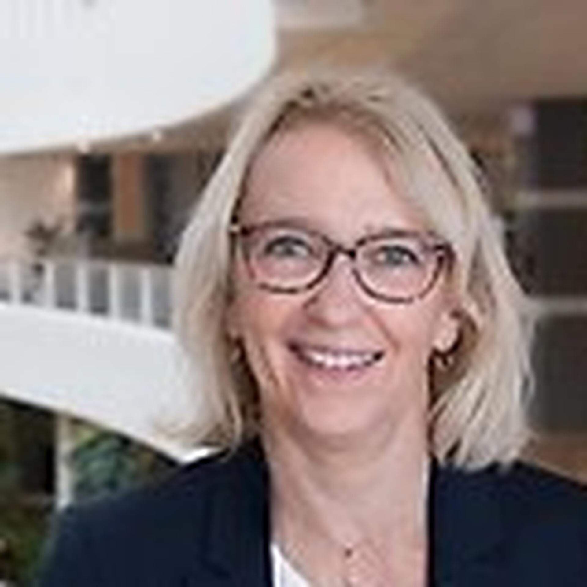 Monica Sonde i kontorsmiljö iklädd vit topp och svart kavaj samt glasögon med mörka bågar