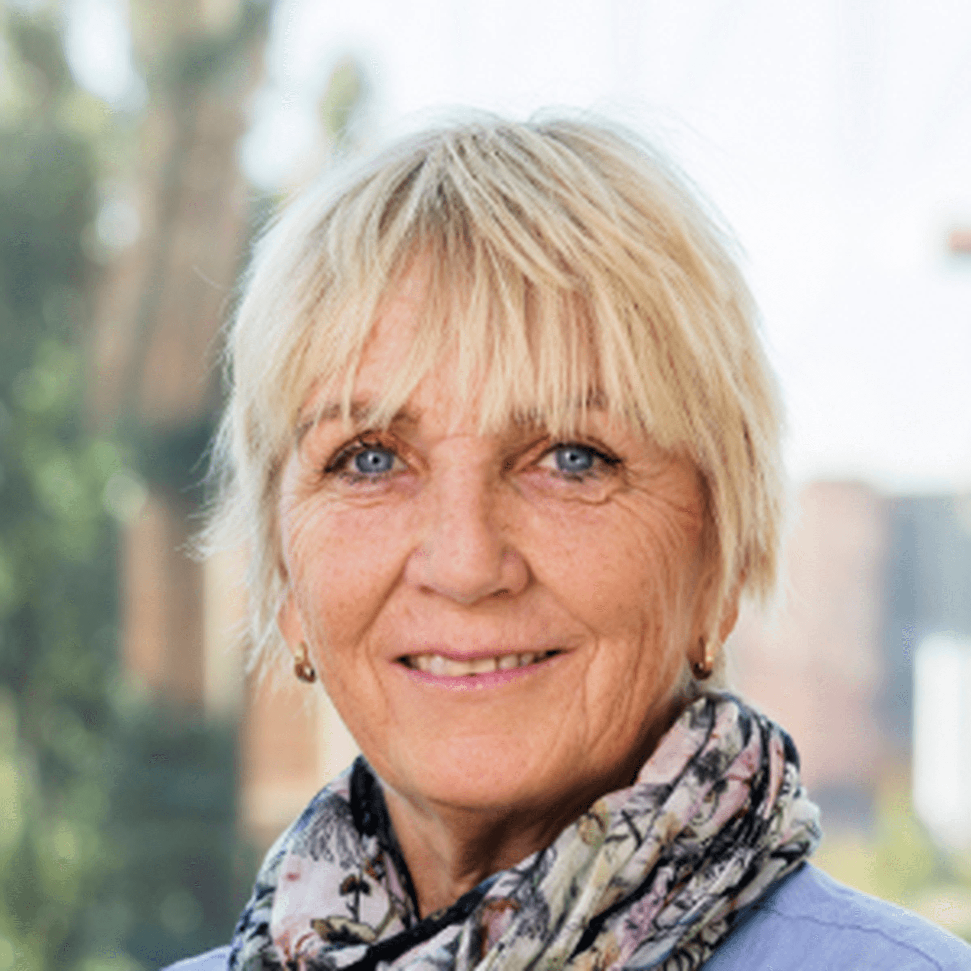 Pia Bergman i ljusblå jacka och blommig sjal. Träd och tegelbyggnader i bakgrunden