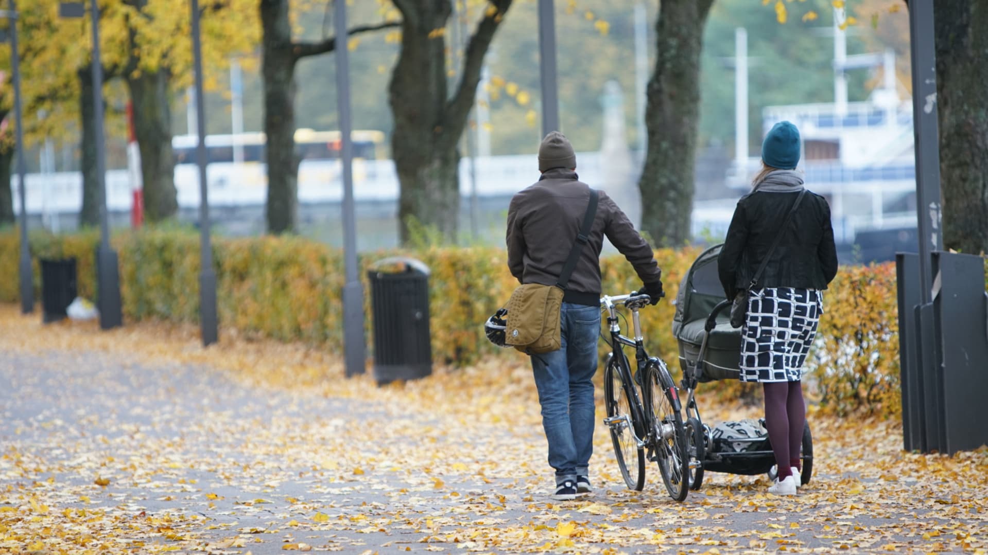 En man leder cykel bredvid en kvinna som går med barnvagn på en gågata fylld av höstlöv.