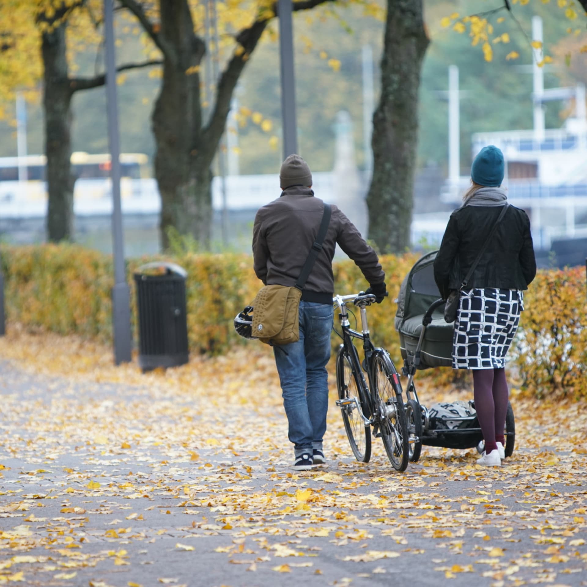 En man leder cykel bredvid en kvinna som går med barnvagn på en gågata fylld av höstlöv.
