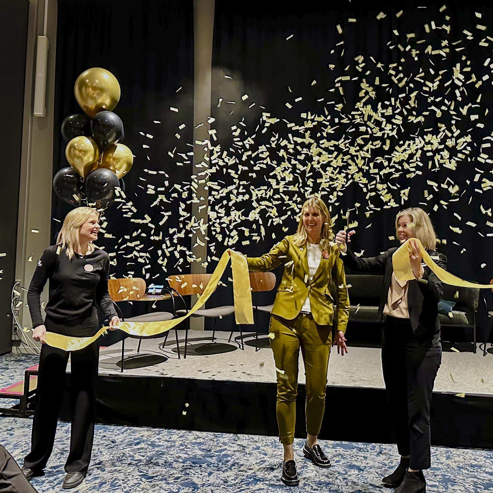 Tre personer håller i ett guldband och det är ballonger och confettiregn