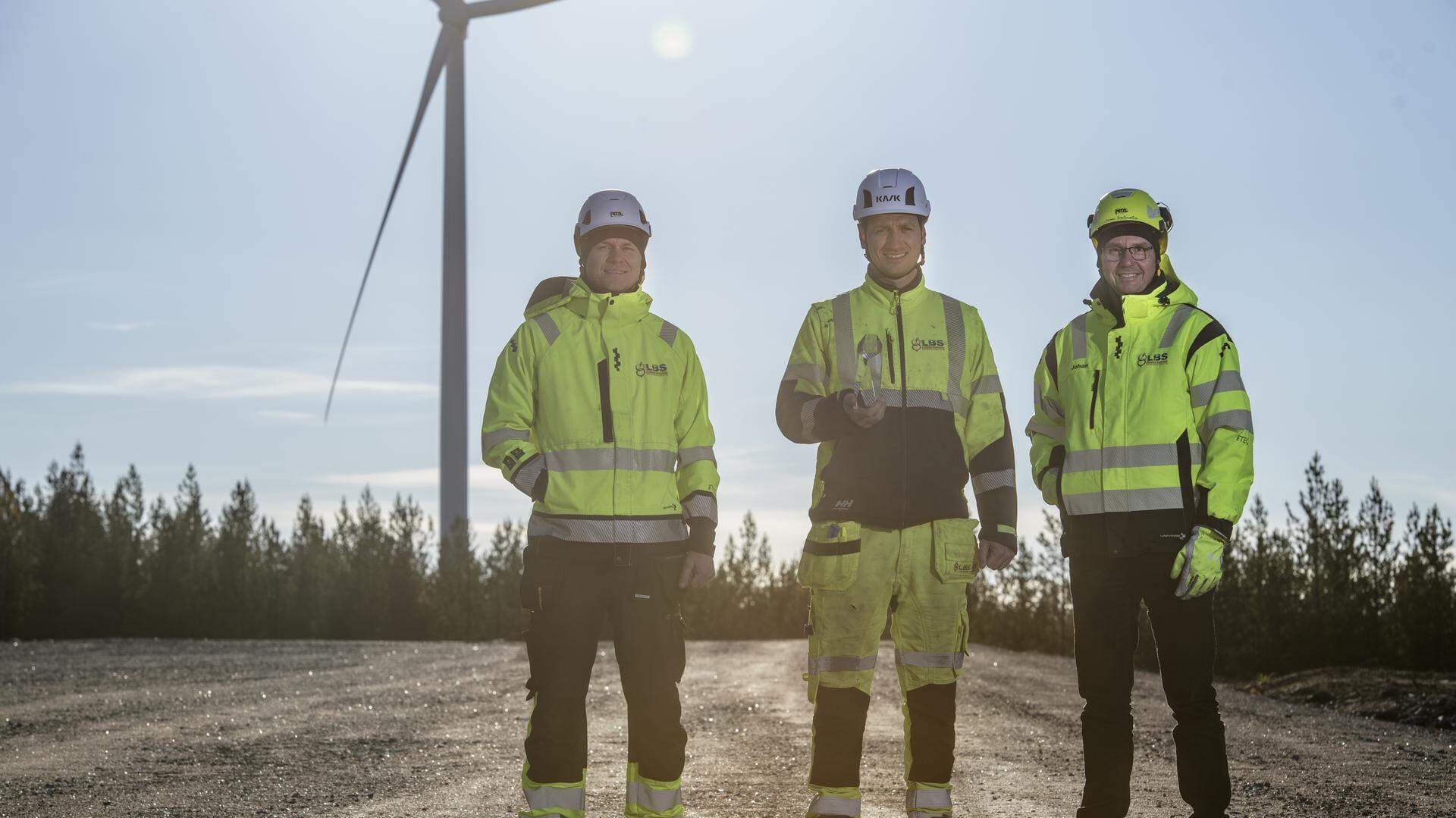 Tre män i arbetskläder framför ett vindkraftverk