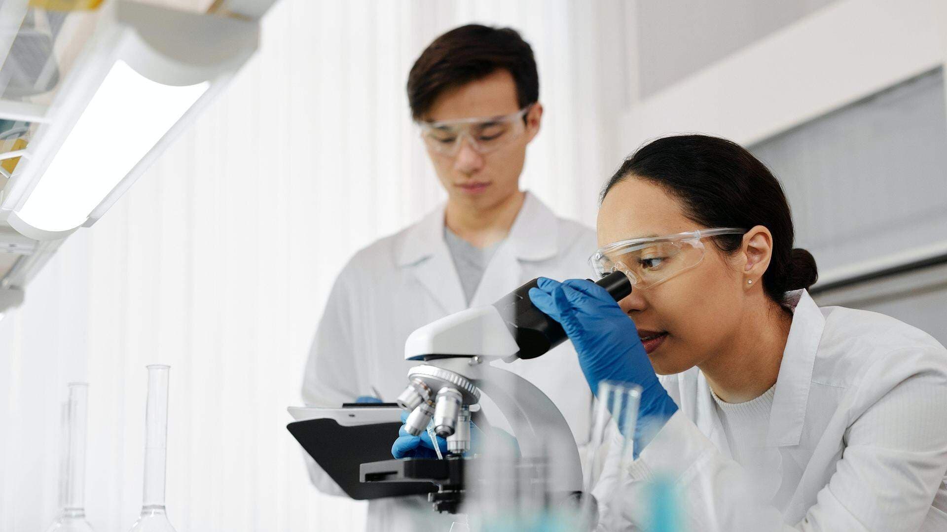 Kvinna som tittar i ett mikroskop och en man som för anteckningar i bakgrunden.