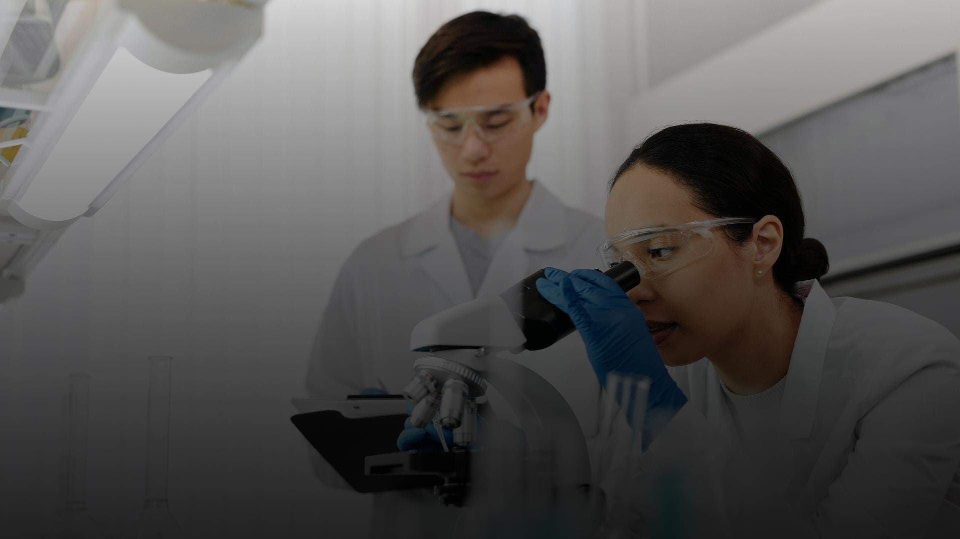 Kvinna som tittar i ett mikroskop och en man som för anteckningar i bakgrunden.
